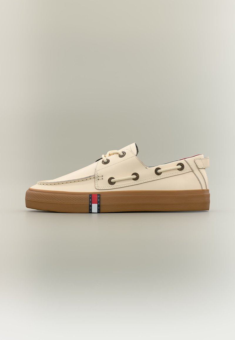 Chaussure bateau en cuir beige basse avec semelle en caoutchouc marron, détails de lacets à travers des œillets métalliques et une petite étiquette logo rouge, blanc et bleu sur la semelle.