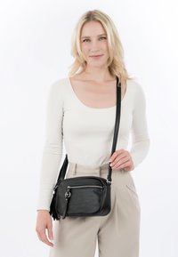 Frau mit blonden Haaren, die ein weißes Langarmshirt und beigefarbene Hose trägt, hält eine schwarze Ledertasche mit einem Reißverschlussfach vorne.