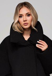 Černá péřová bunda s vysokým límcem, s hladkou texturou a unikátním designem záhybů. Modelka ukazuje bundu s rukama umístěnýma na límci.