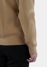 Beige geribde trui met lange mouwen en een hoge hals, met een klein logo in het rood aan de zoom. Het materiaal lijkt gebreid en heeft een gestructureerde textuur.