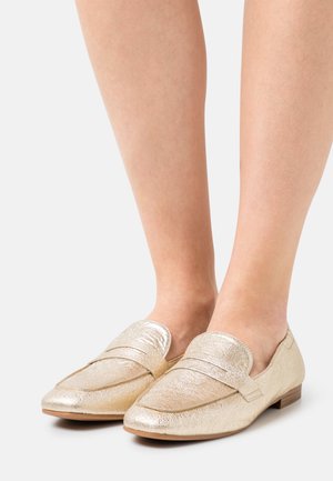 Gambe con mocassini in oro metallizzato dalla texture dettagliata e tacchi bassi in legno su sfondo bianco.