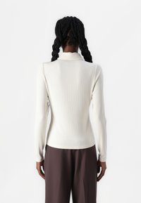 Ribbet, langermet topp i off-white. Har høy hals og tettsittende silhuett. Materialet ser mykt og elastisk ut. Bæres med brune bukser.