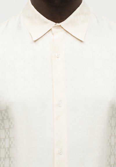 Άτομο με σκούρο δέρμα που φοράει ένα λευκό πουκάμισο με κουμπιά, με διακριτικό καρό σχέδιο και μυτερό γιακά.