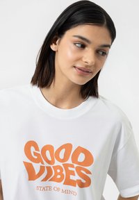 Witte katoen T-shirt met een oranje grafische print waarop "GOOD VIBES" staat en daaronder de tekst "STATE OF MIND". Ronde halslijn.