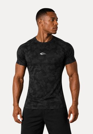 Muskuløs mand, der kigger til højre, iført en tætsiddende sort camo kortærmet træningsshirt med et lille hvidt logo på brystet.