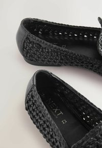 Zapatos planos negros con material tejido y texturizado, y un ribete de cuero negro brillante. Cuentan con una punta redondeada y una plantilla acolchada.