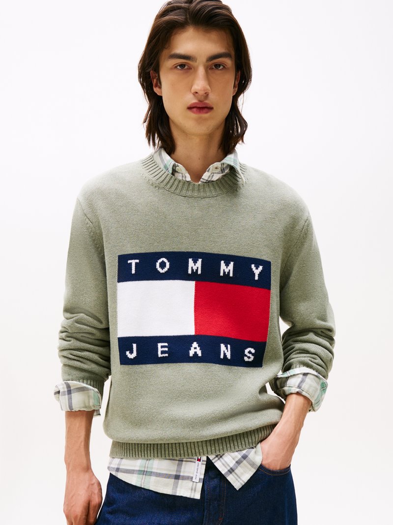 Zeleni pleteni pulover z rebrastim izrezom, ki ima čez prsa velik pravokotni logotip "TOMMY JEANS" v modri, rdeči in beli barvi. Obložen je s srajco s karirastim vzorcem.