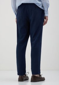 Pantaloni in lino blu navy con una vestibilità rilassata, caratterizzati da un dettaglio a bottone sul retro e gambe leggermente affusolate. Indossati con scarpe marroni.