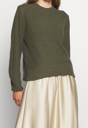 Woman wearing an olive green ribbed knit sweater with long sleeves and a cream-colored satin skirt against a plain background.