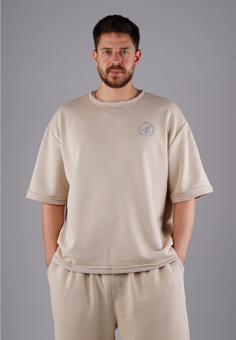 Beige übergroßes T-Shirt mit rundem Ausschnitt, umgeschlagenen Ärmeln und strukturiertem Saum. Verfügt über ein dezentes Logo auf der linken Brust. Weicher, glatter Stoff.