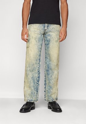 Mężczyzna ubrany w jasnoniebieskie proste jeansy acid-wash, czarne buty i czarną koszulę, stojący na tle jednolitego jasnego tła.