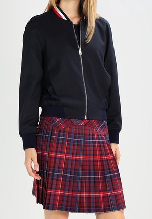 Personne portant une veste noire zippée avec un col rouge, blanc et bleu et une jupe plissée à carreaux rouges et bleus.