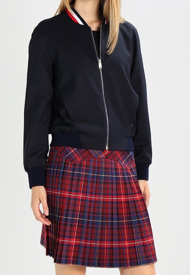 Personne portant une veste noire zippée avec un col rouge, blanc et bleu et une jupe plissée à carreaux rouges et bleus.