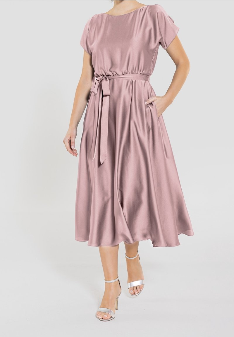 Femme portant une robe midi en satin mauve avec des manches courtes, une ceinture à la taille, des poches latérales et des sandales argentées à talons hauts, marchant vers l'avant.