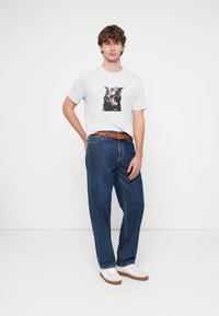 Λευκό βαμβακερό t-shirt με μαύρο σχέδιο σκύλου, σε συνδυασμό με χαλαρά μπλε τζιν και λευκά αθλητικά παπούτσια με καφέ σόλες, συνοδευόμενο από καφέ ζώνη.