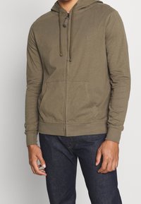 Sudadera con capucha y cremallera de color verde oliva, fabricada con tela suave, con bolsillo tipo canguro, capucha ajustable con cordón y puños acanalados. Diseño sencillo.