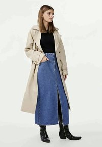 Beige trenchcoat med belted midje, båret over en svart topp og et blått denim maxi-skjørt. Skjørtet har en frontspalte og rett passform.
