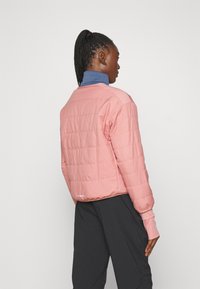 Veste matelassée rose avec une texture lisse, encolure arrondie et poignets ajustés, superposée sur un col roulé bleu, associée à un pantalon noir.