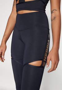 Mustat korkea vyötäröiset legginssit, joissa on leikattu yksityiskohtia reisissä. Sivulla on kultalogo ja kangas on sileä ja joustava.