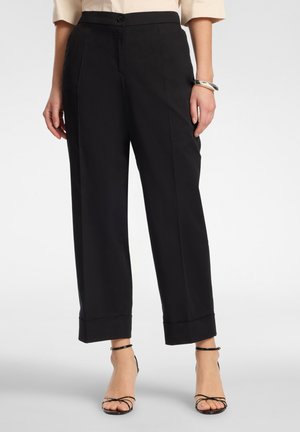 Frau trägt hochgeschnittene schwarze kurze Hosen mit umgeschlagenem Saum und schwarze Riemchen-Sandaletten mit Absatz, steht vor einem schlichten Hintergrund.