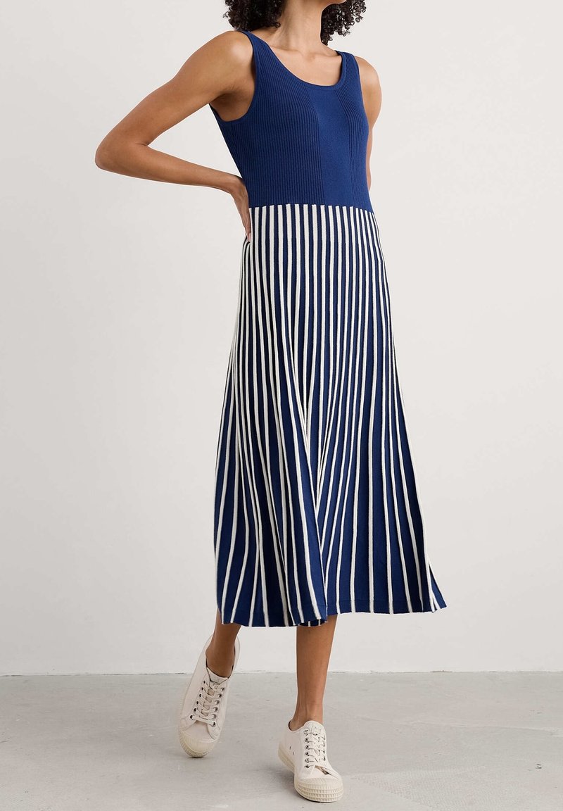 Robe débardeur en maille côtelée bleu marine avec rayures blanches verticales, silhouette évasée en ligne A, portée avec des baskets blanches sur un sol gris.