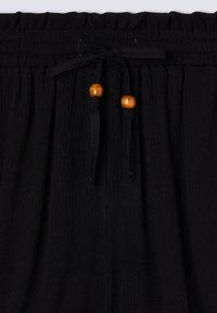 Czarne spodnie z materiału plisowanego z elastyczną talią, wyposażone w sznurek związany drewnianymi koralikami jako akcent.