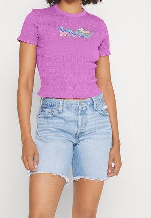 Kvinde iført en tekstureret lilla cropped t-shirt med et farverigt Levi's-logo og lyseblå højtaljede denimshorts med frynset kant.
