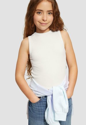 Junges Mädchen mit langen, lockigen Haaren, das ein ärmelloses weißes Oberteil und eine blaue Jeans trägt, mit einem hellblauen Sweatshirt um die Taille gebunden und die Hände in den Taschen.