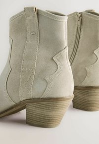 Botas de ante hasta el tobillo en verde claro con costuras decorativas en zigzag y un tacón bloque texturizado. Presentan una cremallera lateral para facilitar su uso.