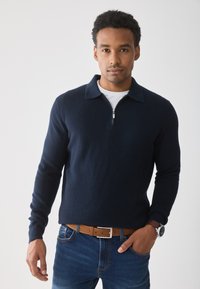 Marineblauwe geribbelde trui met een ritssluiting op de kraag, gedragen over een wit T-shirt, in combinatie met een blauwe spijkerbroek en een bruine riem. Zilveren horloge om de pols.
