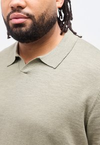 Κοντινό πλάνο ενός ατόμου με γενειάδα και σκούρα μαλλιά σε πλεξίδες, που φοράει ένα ανοιχτοπράσινο πλεκτό πουλόβερ με γιακά, μπροστά σε  απλό φόντο.