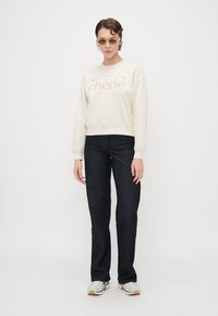 Krem crew-neck sweatshirt med "chérie" grafikk, kombinert med mørke jeans. Modellen har på seg solbriller og lyse sneakers. Minimalistisk design.