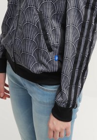 Černá a bílá zipová bunda s geometrickým vzorem, s pevnými černými akcenty na rukávech a kapsách, a s lemovaným límcem.