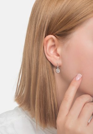 Silberne Tropf-Ohrringe mit floralem Design hängen von einem Ring, präsentiert vor einem hellen Hintergrund, getragen von einer Person mit glattem Haar.