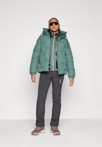 Kvinna som bär en grön pufferjacka, mönstrad zip-up tröja, mörka byxor, rosa vandringsskor och överdimensionerade solglasögon som står mot en vit bakgrund.