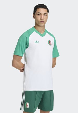 Junger Mann steht mit den Händen hinter dem Rücken, trägt ein weiß-grünes Fußballtrikot von Algerien mit Adidas-Logo und Länderabzeichen.