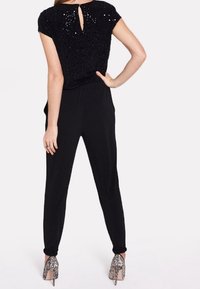 Top negro con lentejuelas y mangas cortas, con un escote en la espalda; combinado con pantalones negros ajustados y tacones altos plateados brillantes.