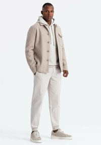 Veste en laine gris clair avec des boutons, superposée à un sweat à capuche léger, associée à un pantalon clair et des baskets beiges dans un cadre neutre.