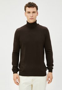 Ciemnobrązowy sweter z golfem wykonany z miękkiego materiału dzianinowego, z długimi rękawami i ściągaczami. Model nosi jasne spodnie.