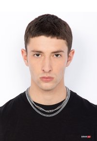 Collane di catena argentata sovrapposte a una maglietta nera, con collo rotondo e un piccolo logo sul petto. Sfondo semplice.