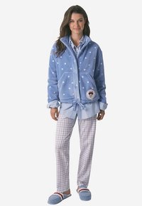 Světlá modrá fleecová bunda na zip s bílými vzory, doplněná modro-bílou kostkovanou košilí a světlými kostkovanými kalhotami. Modré pantofle doplňují vzhled.