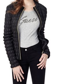 Giacca nera trapuntata con zip dorata, indossata sopra una t-shirt grigia con scritta nera in perline, abbinata a pantaloni neri.