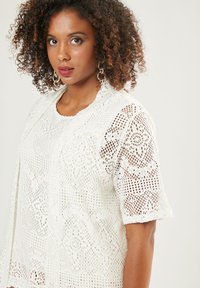 Top bianco all'uncinetto con maniche corte, caratterizzato da motivi intricati e trama aperta. Nota per la sua texture simile al pizzo e il design leggero.