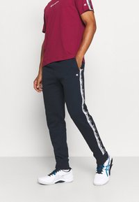 Mörkröd t-shirt med kort ärm och logotyp; marinblå sweatpants med vita sidostrecken och logotyp; vita sportskor med blå detaljer.