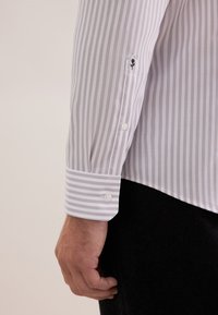 Camicia a maniche lunghe bianca con strisce verticali grigio chiaro; presenta un polsino con bottoni, dettaglio di fiore ricamato e una texture del tessuto liscia.