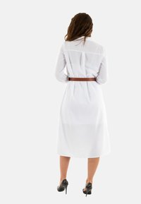 Robe blanche avec des manches longues, taille ceinturée et longueur midi. Tissu lisse et détail de col classique. Portée avec des chaussures à talons hauts.