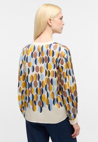 Blusa a maniche lunghe realizzata in tessuto leggero, caratterizzata da un motivo astratto di cerchi sovrapposti nei colori blu, giallo, marrone e crema.