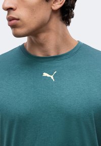 Νέος άνδρας που φοράει πετρόλ μπλουζάκι Puma με λευκό λογότυπο Puma κεντραρισμένο κάτω από το λαιμό, κοιτάζει αριστερά σε απλό φόντο.
