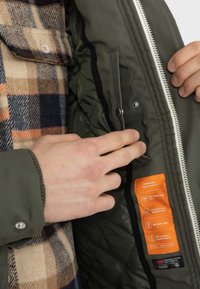 Grüne Außenjacke mit gestepptem Futter, praktischen Reißverschlusstaschen und einem orangen Etikett, das die Leistungsmerkmale hervorhebt.