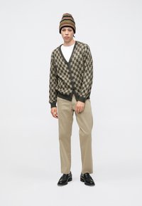 Stickad kofta med schackrutigt mönster i mörkbrunt och beige, kombinerad med beige byxor och en randig mössa. Svarta skor.